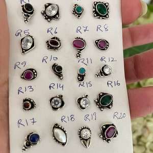 May include: Twenty nose rings with various colored gemstones and settings. The rings are arranged in rows on a white card with labels for each ring.