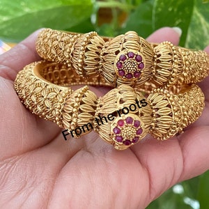 May include: Two gold bangles with intricate floral designs and red gemstone accents. The bangles are stacked on top of each other and feature a raised, circular design with a cluster of red gemstones in the center.