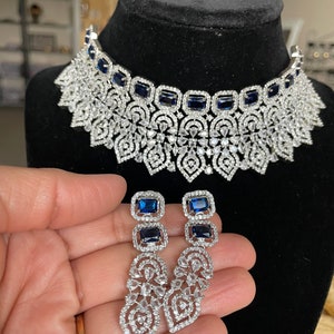 May include: A silver choker necklace with a blue stone design and matching dangle earrings. The necklace has a large, intricate design with many small stones. The earrings are long and have a similar design to the necklace.