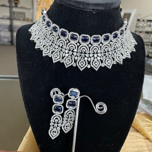 May include: A silver necklace and earrings set featuring a delicate floral design and blue gemstones. The necklace is a choker style with a large, intricate pendant. The earrings are long and dangle.