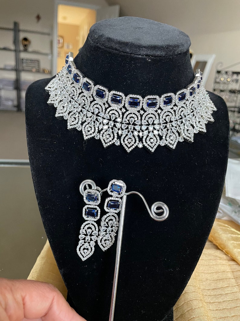 May include: A silver necklace and earrings set featuring a delicate floral design and blue gemstones. The necklace is a choker style with a large, intricate pendant. The earrings are long and dangle.