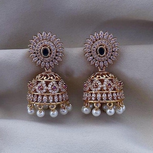 May include: A pair of gold-toned, ornate earrings. Each earring features a floral-shaped top with a dark center stone, a bell-shaped body with intricate detailing, and dangling white pearls. The earrings are set against a neutral background.