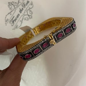 May include: Gold bangle bracelet with a square shape and a black and white design. The bracelet is decorated with red gemstones and small diamonds.