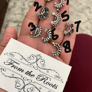 May include: Eight silver nose rings with intricate designs, displayed on a hand. The rings are numbered 1 through 8. The card in the background reads "From the Roots".