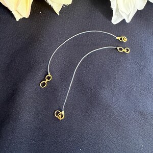 May include: Three gold-colored chain extenders with clear, thin cords. Each extender has a small, circular clasp at both ends. The extenders are arranged on a dark background, with white and yellow floral accents in the upper corners.