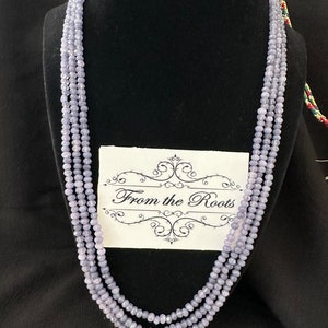 May include: A multi-strand necklace made of light purple beads. The necklace is displayed on a black velvet mannequin bust. The tag on the necklace reads "From the Roots".