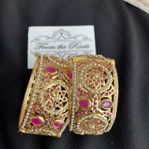 May include: Two gold bangles with intricate designs, featuring pink gemstones and white pearls. The bangles are stacked on top of each other and have a delicate, ornate look. The text "From the Roots" is visible on a white card behind the bangles.