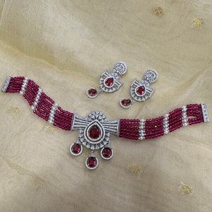 May include: A red and white beaded choker necklace with a silver pendant featuring a large red gemstone surrounded by smaller white gemstones. Matching earrings with a similar design are also included.