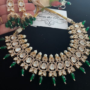 May include: A gold necklace with a large, square-shaped pendant adorned with clear stones and surrounded by smaller stones. The pendant is attached to a chain with small white pearls and green beads. The necklace is accompanied by a pair of matching earrings with a similar design.