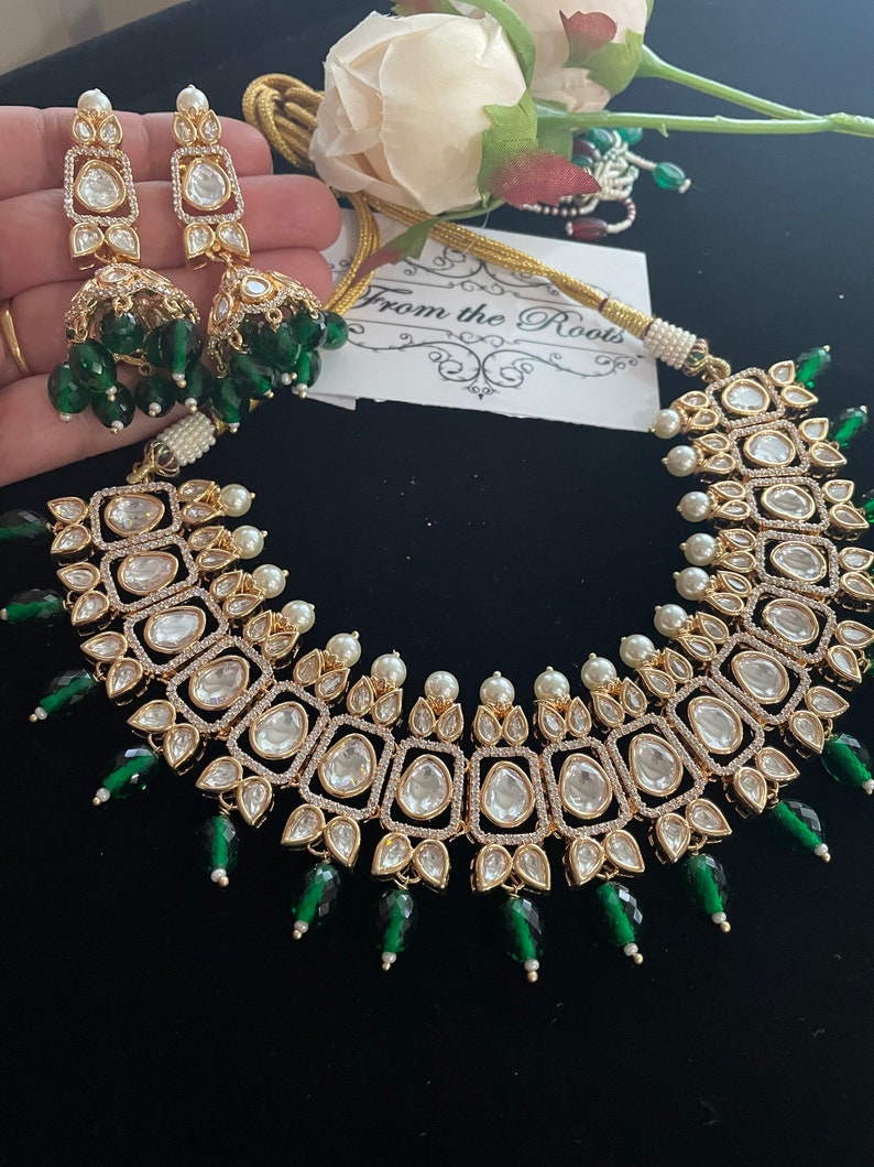 May include: A gold necklace with a large, square-shaped pendant adorned with clear stones and surrounded by smaller stones. The pendant is attached to a chain with small white pearls and green beads. The necklace is accompanied by a pair of matching earrings with a similar design.