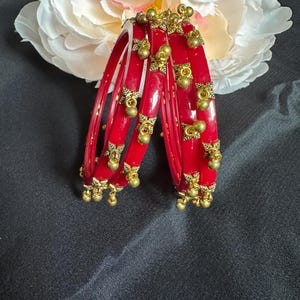 May include: Set of red glass bangles with gold accents and dangling bells. The bangles are stacked on top of each other.