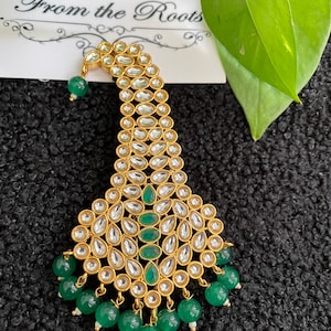 May include: A gold hair ornament with green and white stones. The ornament is shaped like a crescent moon and has a beaded fringe.