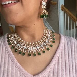 May include: A gold necklace with a green and white design. The necklace has a pendant with a square shape and is decorated with small, round stones. The necklace is worn by a person with a pink shirt.