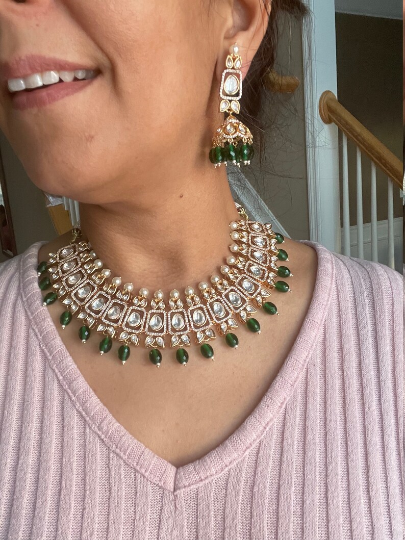 May include: A gold necklace with a green and white design. The necklace has a pendant with a square shape and is decorated with small, round stones. The necklace is worn by a person with a pink shirt.