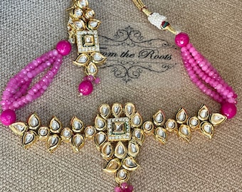 May include: A gold necklace with a pink beaded chain and a large, ornate pendant with a square center and teardrop-shaped stones. The pendant is attached to a smaller, matching pendant with a similar design. The necklace is accompanied by a pair of matching earrings.