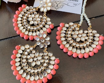 May include: A pair of large, gold-tone earrings with a circular design. The earrings are adorned with clear stones and coral-colored beads. The earrings are hanging from a white string of pearls.