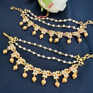 May include: A pair of gold-tone ear cuffs with pearl chains and dangling floral accents. The floral accents are adorned with clear and red gemstones.