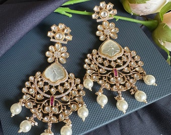 May include: A pair of gold earrings with white pearl accents. The earrings feature a floral design with a white stone center and a delicate, intricate pattern.