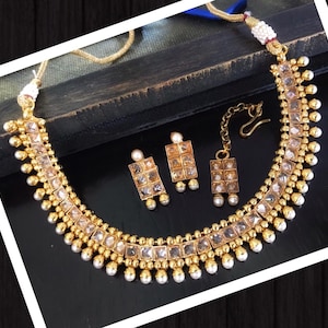 May include: A gold necklace with a row of square-shaped stones and pearl accents. Matching earrings and a nose ring are also included.