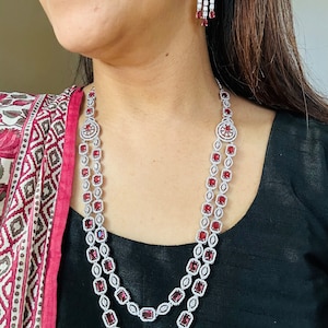 Puede incluir: Un collar de plata con una doble hilera de piedras rojas y transparentes. El collar tiene un diseño delicado con pequeñas piedras de forma cuadrada. El collar se combina con pendientes a juego con un diseño similar.