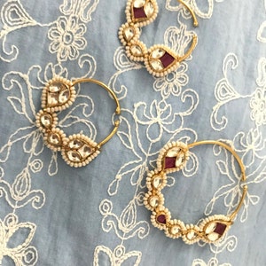 May include: Three gold nose rings with white pearl and red gemstone accents. The rings are on a white and blue patterned fabric.