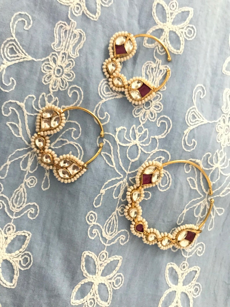 May include: Three gold nose rings with white pearl and red gemstone accents. The rings are on a white and blue patterned fabric.