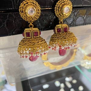 May include: Gold-toned earrings with a floral design and a clear stone at the top. The earrings feature red square stones and a bell-shaped bottom with white pearls and red teardrop-shaped stones.