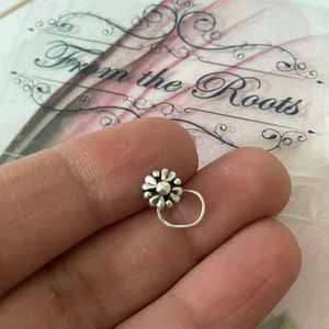 May include: A silver nose ring with a flower design. The nose ring has a small, detailed flower at the top and a curved wire that goes into the nose. The background includes the text "From the Roots".