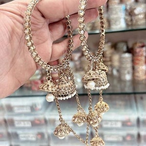 May include: A pair of gold-tone bangles with dangling charms. The charms are made of metal and have a pearl-like finish. The bangles are adorned with clear rhinestones.