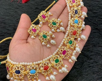 May include: A gold-toned necklace and earring set featuring colorful gemstones and faux pearls. The necklace and earrings have a floral design with red, blue, green, and pink stones. The set is displayed on a hand.