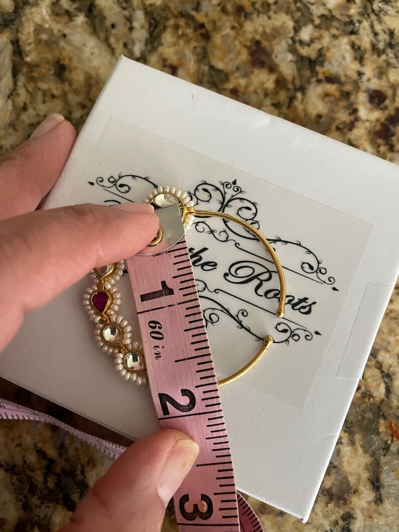 May include: A gold nose ring with white pearls and pink stones, measuring 1.6 inches in diameter. The ring is on a white box with the words "The Roots" printed on it.