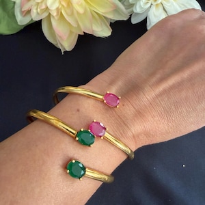 May include: Gold-toned metal bracelets, each featuring a single oval gemstone. One bracelet has a ruby-colored stone, while the other two have emerald-colored stones. The bracelets are displayed on a wrist against a dark background.