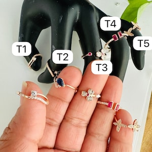 May include: A set of ten gold-tone rings with various designs, including a flower, a butterfly, and a square. The rings are set with clear and blue gemstones. The rings are labeled T1 through T5.