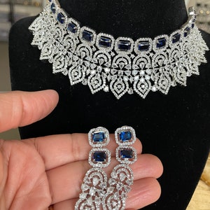 May include: A silver and blue statement necklace and matching earrings. The necklace features a delicate filigree design with blue square-shaped stones. The earrings are long and dangle, with a similar filigree design and blue stones.