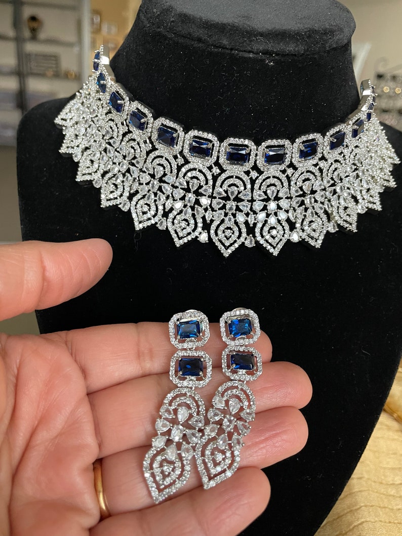 May include: A silver and blue statement necklace and matching earrings. The necklace features a delicate filigree design with blue square-shaped stones. The earrings are long and dangle, with a similar filigree design and blue stones.