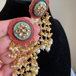 May include: A pair of long, dangling earrings with a round, red and green enamel design. The earrings are adorned with small, white pearls and gold accents.