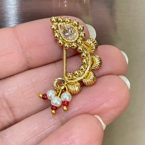May include: A gold nose ring with a clear stone and a dangling charm of white pearls and red beads.