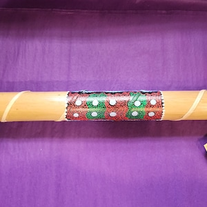 May include: A cylindrical didgeridoo made of bamboo, featuring a central band with a painted design of red, green, and white dots. The bamboo is wrapped with thin strips, and a tag hangs from the end. The background is a purple surface.