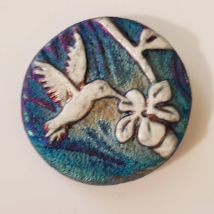 May include: A round, ceramic hummingbird art piece. The hummingbird is white with a red outline, hovering near a white flower. The background is a blend of blue, teal, and purple hues, creating a textured effect. The art piece is a decorative item.