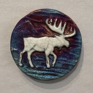 May include: A round, decorative ceramic piece featuring a white moose against a backdrop of swirling blues, purples, and bronze hues. The moose is the focal point, with detailed antlers and a textured surface. The piece has a rustic, handcrafted appearance.