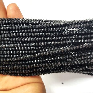 May include: Close-up of multiple strands of small, faceted black beads. The beads are strung together, creating a dense, textured surface. The beads are held in a hand, with a white background.