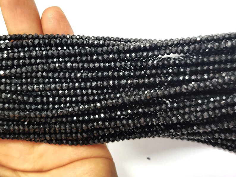 May include: Close-up of multiple strands of small, faceted black beads. The beads are strung together, creating a dense, textured surface. The beads are held in a hand, with a white background.