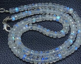 Op de afbeelding: Een ketting bestaande uit talloze kleine, gefacetteerde, doorschijnende maansteen kralen. De kralen vertonen een subtiele blauwe adularescentie of glans wanneer er licht op hun oppervlak valt. De streng is bevestigd met een zilverkleurige metalen karabijnsluiting.
