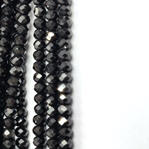 May include: Close-up of multiple strands of black faceted beads. The beads are small, round, and have a shiny, reflective surface. The beads are strung together, creating a vertical pattern against a white background. The overall effect is one of elegance and sophistication.