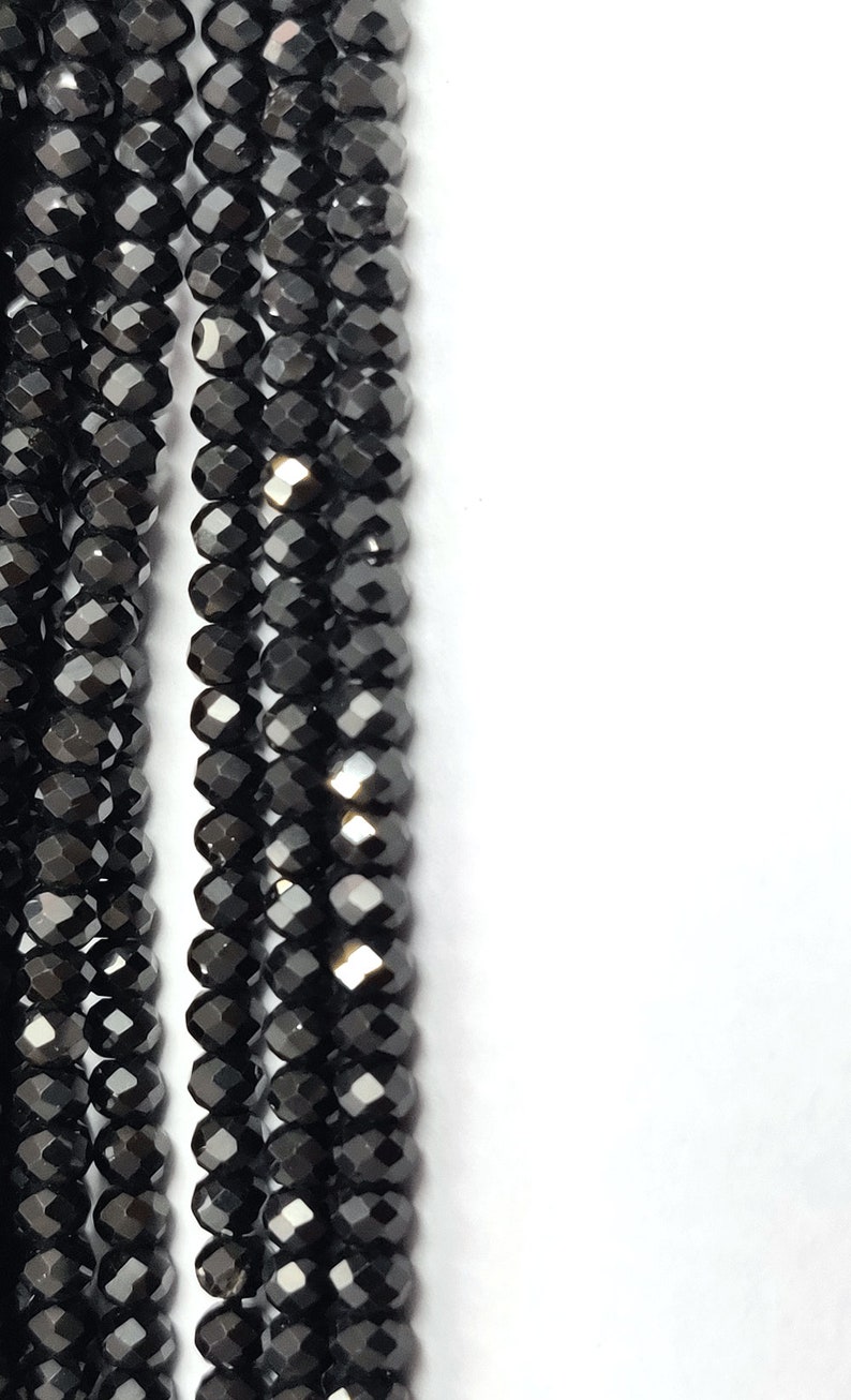 May include: Close-up of multiple strands of black faceted beads. The beads are small, round, and have a shiny, reflective surface. The beads are strung together, creating a vertical pattern against a white background. The overall effect is one of elegance and sophistication.