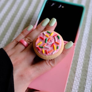 May include: A circular phone grip featuring a tan cookie base topped with pink frosting and multi-colored sprinkles. The accessory is attached to a pink smartphone case, and a solid pink ring is worn on the finger next to the phone grip.