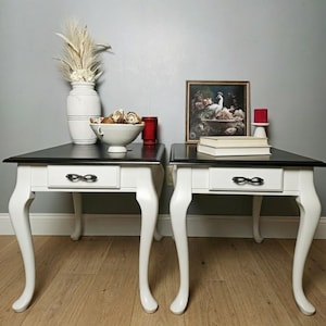 May include: A pair of side tables with black tops and white bases. Each table has a single drawer with a decorative handle and curved legs. One table has a white vase with dried flowers, a bowl, and a red glass. The other table has a framed picture and books.
