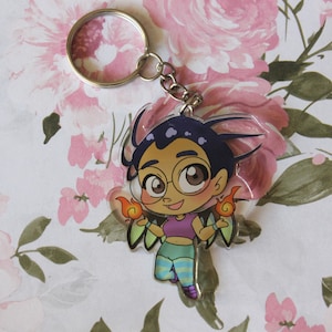May include: A clear acrylic keychain featuring a cartoon character with purple hair, glasses, and wings. The character is holding two flames in each hand. The keychain is on a white background with pink flowers.