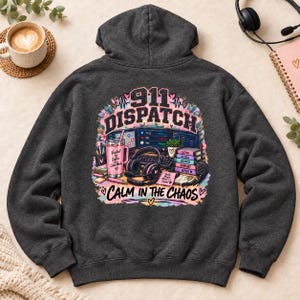 May include: A dark grey hooded sweatshirt featuring a colourful graphic on the back. The design includes the text 911 DISPATCH and CALM IN THE CHAOS, along with illustrations of a computer setup, a pink coffee mug, headphones, and books.