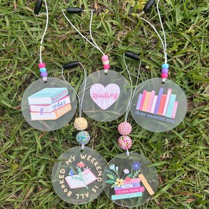 May include: Five clear acrylic car charms with various book-themed designs and text. One charm says "Bookish Vibes Only," another says "In My Reading Era," and a third says "In My Reading Era." The other two charms say "Booked My Weekend Is All" and "Just One More Chapter."